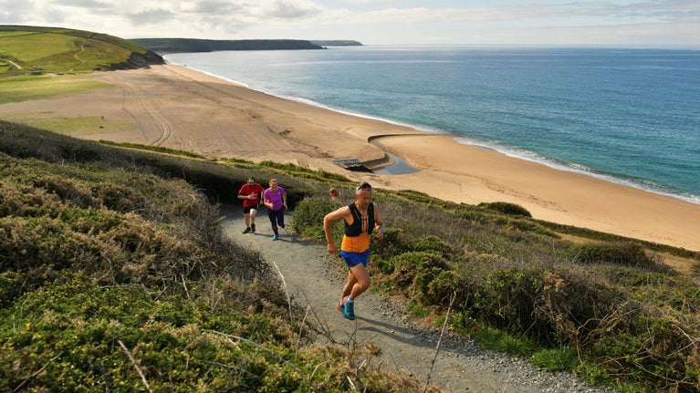 Runners running up a coast path with a beach behind and to the left of them